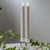 Bézs LED vacsoragyertyák dekor pohárban, meleg fényű lánggal, természetes zöld díszítéssel és elegáns hangulatban elrendezve.